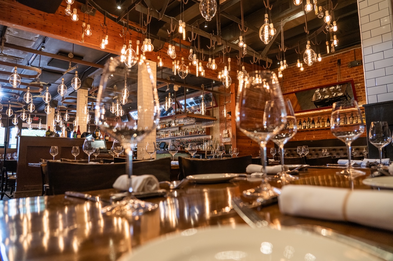 The dining room at Little Alley Steak Roswell — dozens of oversized Edison bulbs hanging from blackened steel above set tables, with the back bar and its bottle shelves beyond exposed brick and reclaimed wood